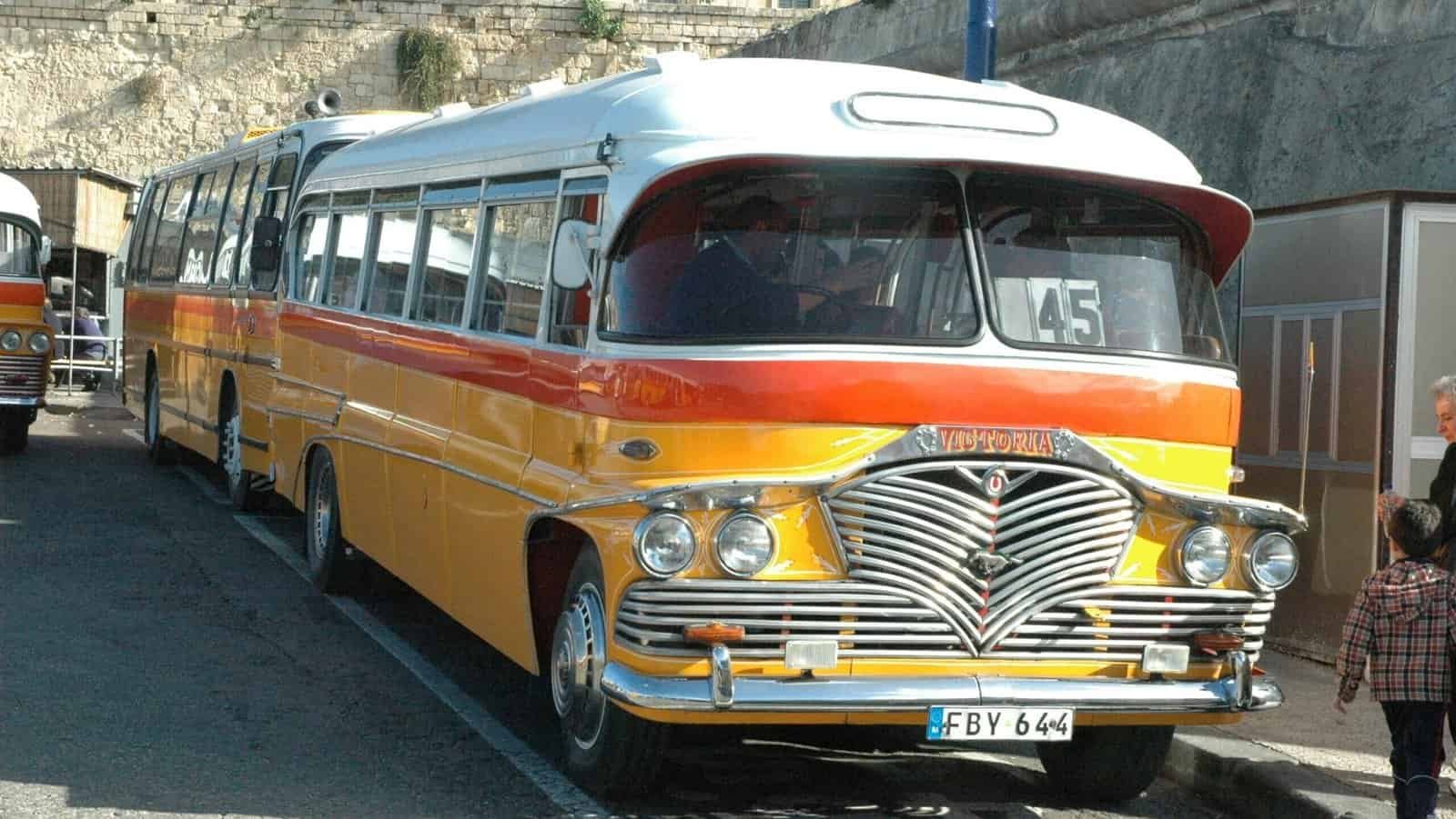 Traditional orange and yellow Malta bus parked next to a wall.