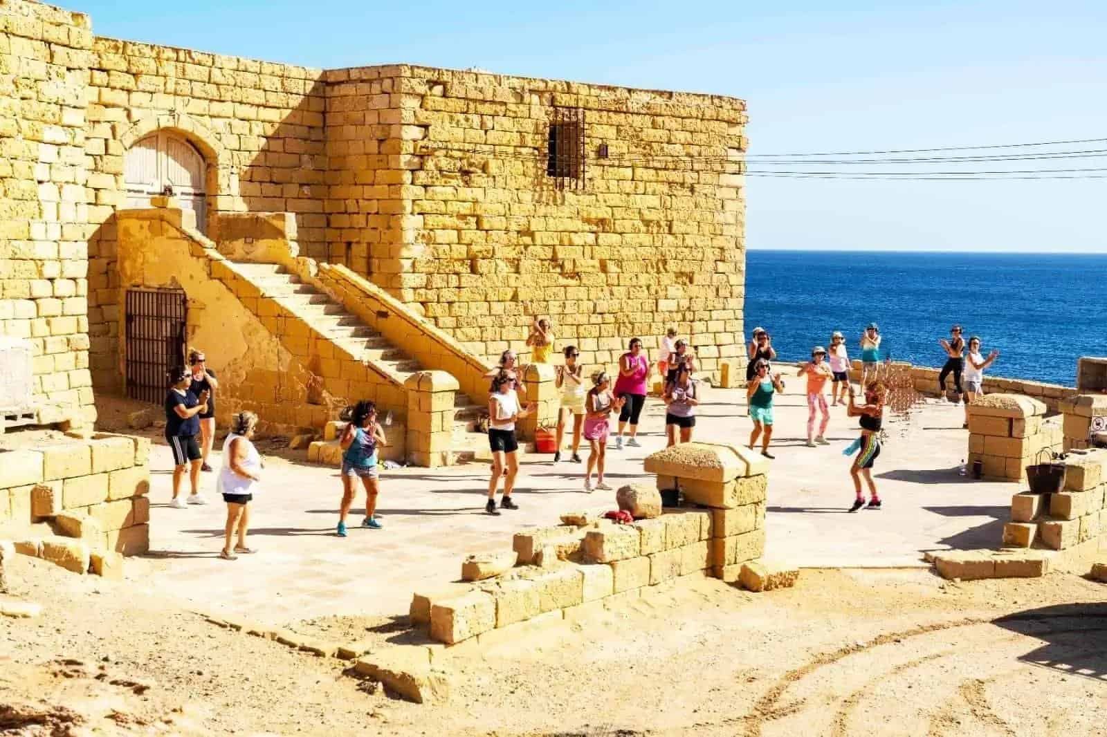 A group of people exercising in front of a limestone fort, overlooking the sea.