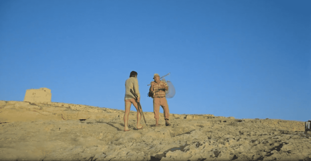 A man walks up a bare hill, another is walking out of sight to the right, carrying a long pole with fishing baskets.