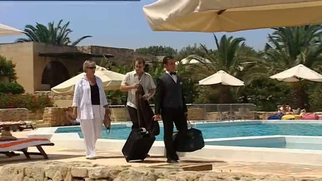 Two men and a woman walking by a hotel swimming pool, dragging suitcases.