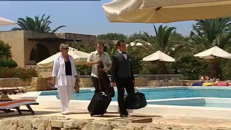 Two men and a woman walking by a hotel swimming pool, dragging suitcases.