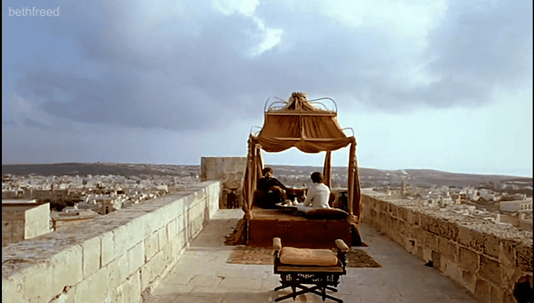 Two men sit on a rooftop, on low chairs under a red and gold canopy.