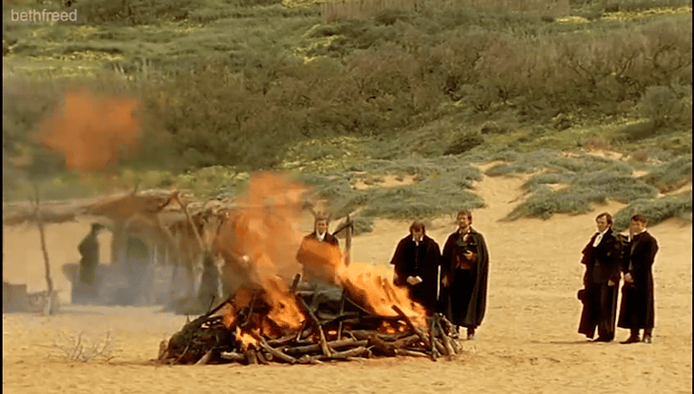 A group of men gather beside a funeral pyre on a sandy beach.