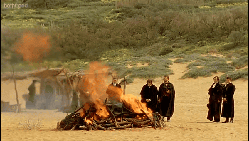 A group of men gather beside a funeral pyre on a sandy beach.