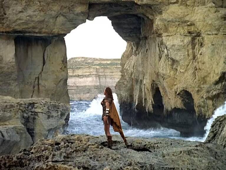 A man, dressed as an ancient greek, stands with his legs and arms wide, in front of the Azure window.