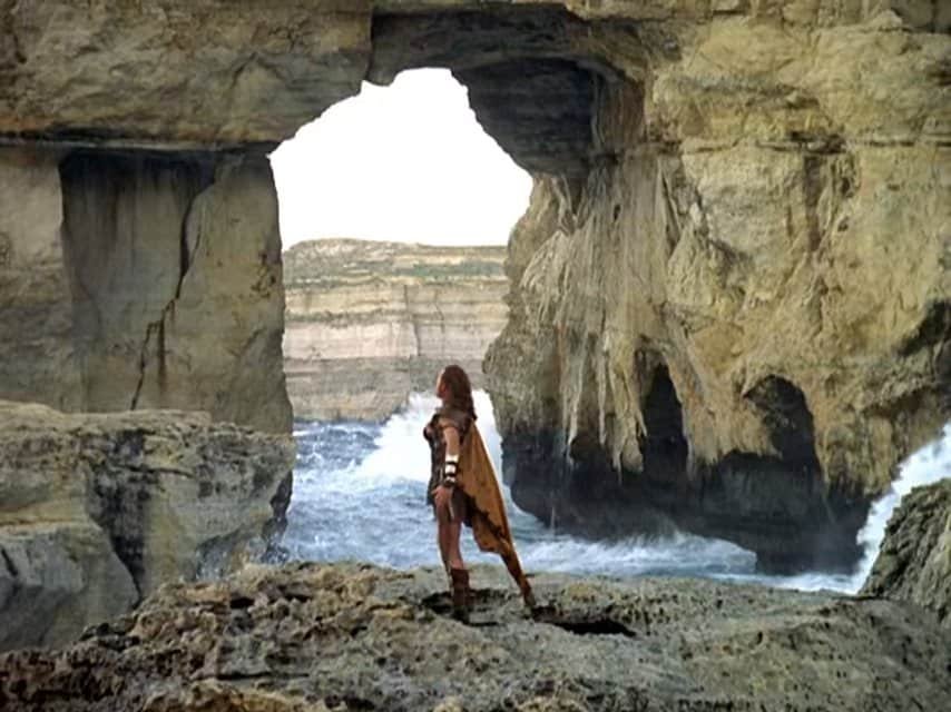 A man, dressed as an ancient greek, stands with his legs and arms wide, in front of the Azure window.