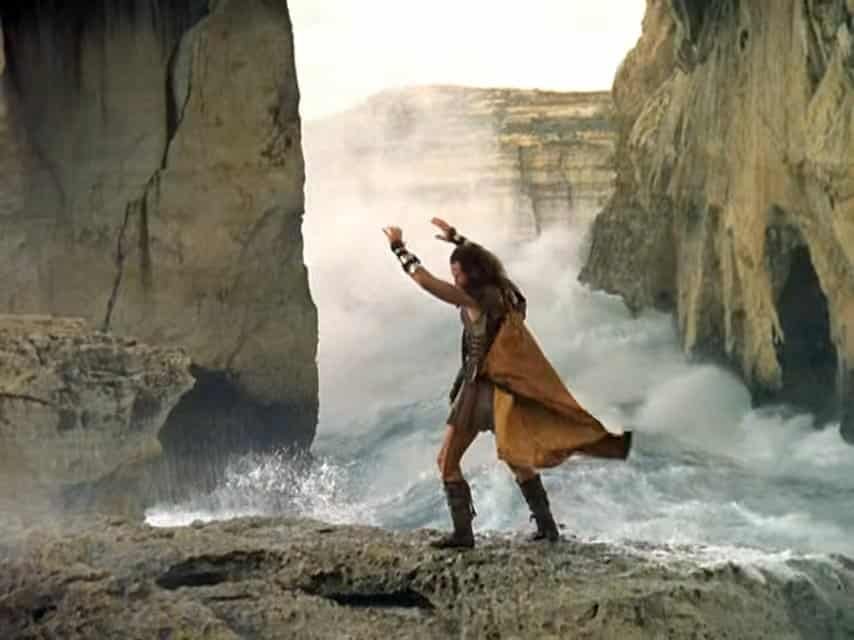 A man, dressed as an ancient greek, stands with his legs and arms wide, on the sea shore.