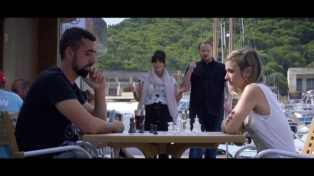 A man and woman play chess on an outdoor table, while a man and woman look on.