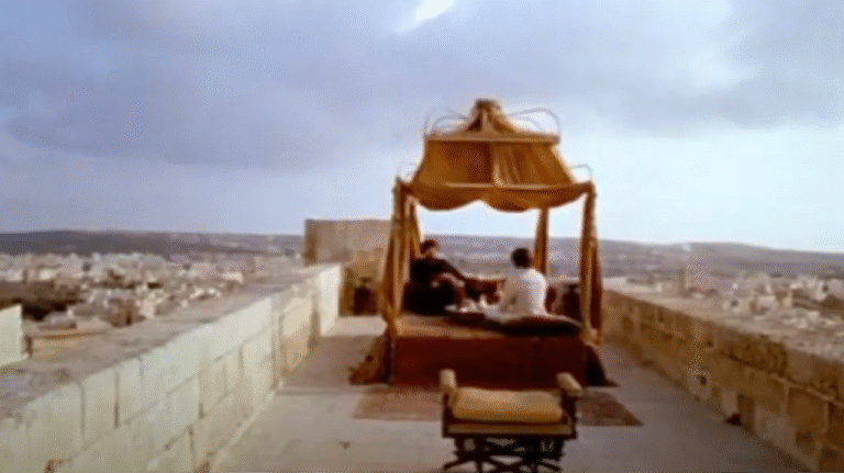 Two men sit on a rooftop, on low chairs under a red and gold canopy.