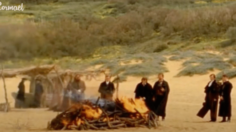 A group of men gather beside a funeral pyre on a sandy beach.