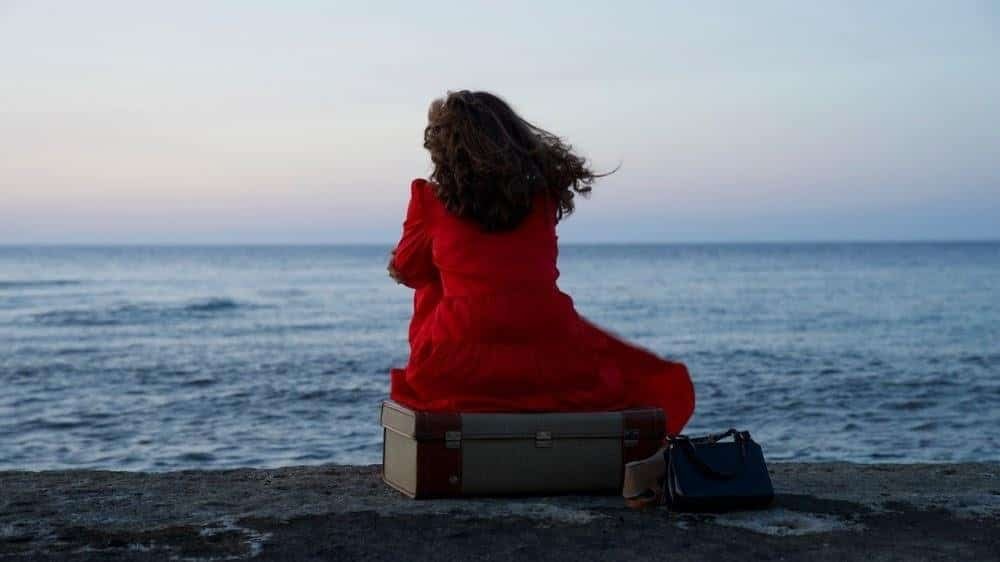 A woman with long brown hair, in a flowing red dress sits on the sea shore, with her back to the camera.