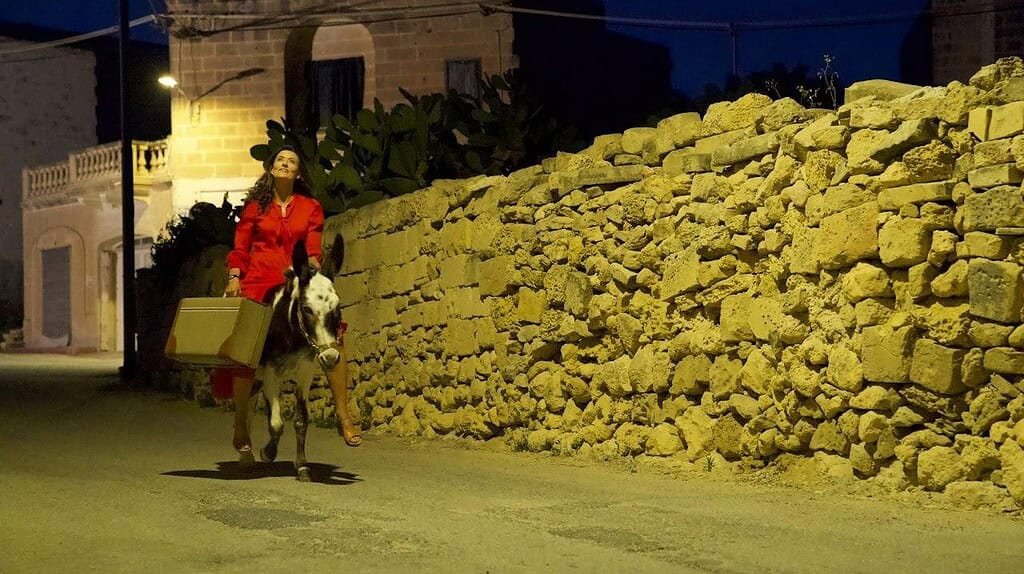 A woman in a flowing red dress rides a donkey on a country lane, next to a rubble wall.