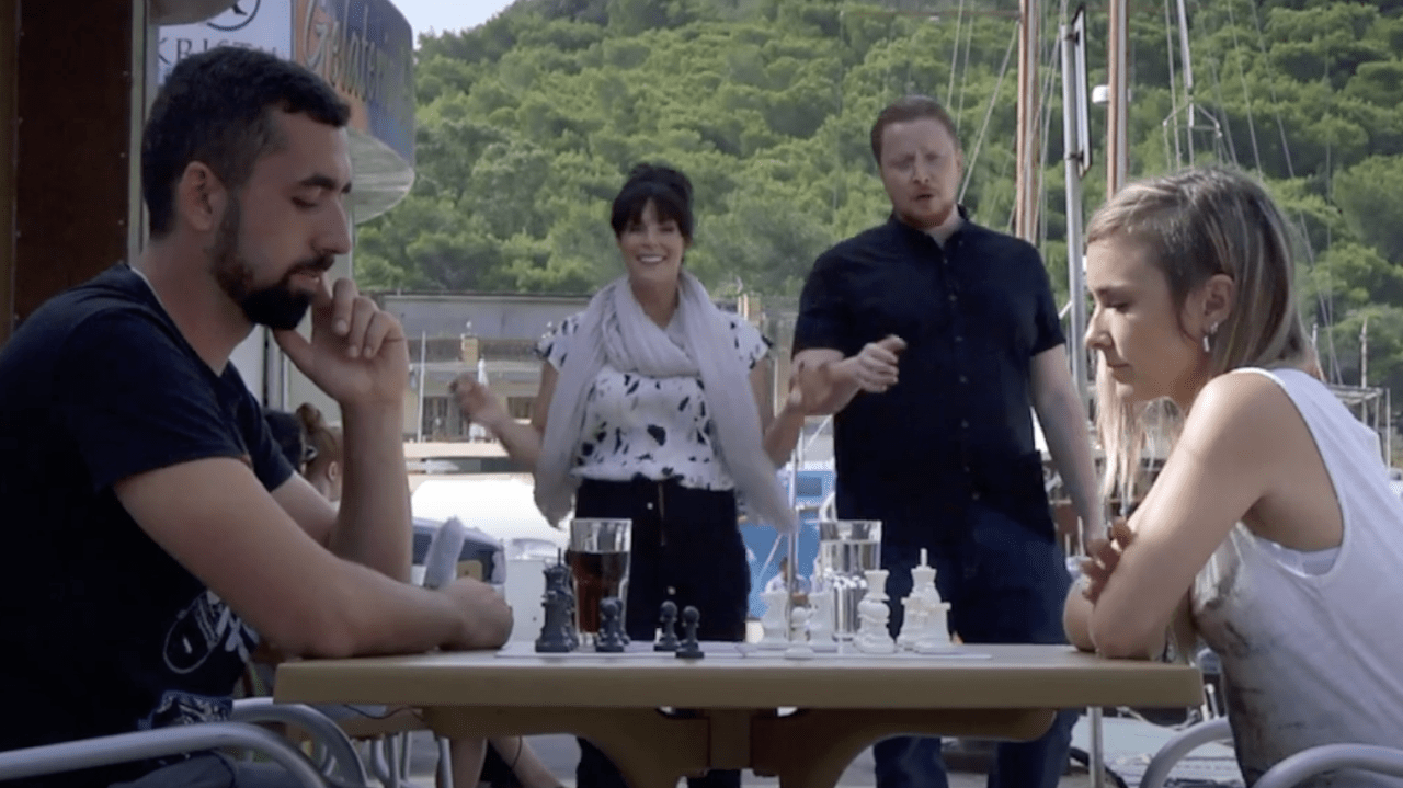 A man and woman play chess on an outdoor table, while a man and woman look on.