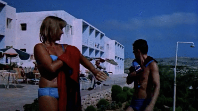A man and woman, dressed in swimming costumes, towel off in front of a two-storey, white, hotel.