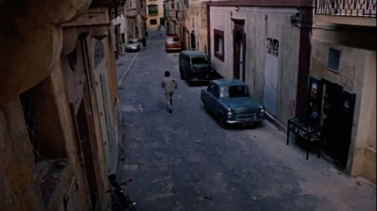 A man, dressed in a tan suit, walks quickly down a narrow road.