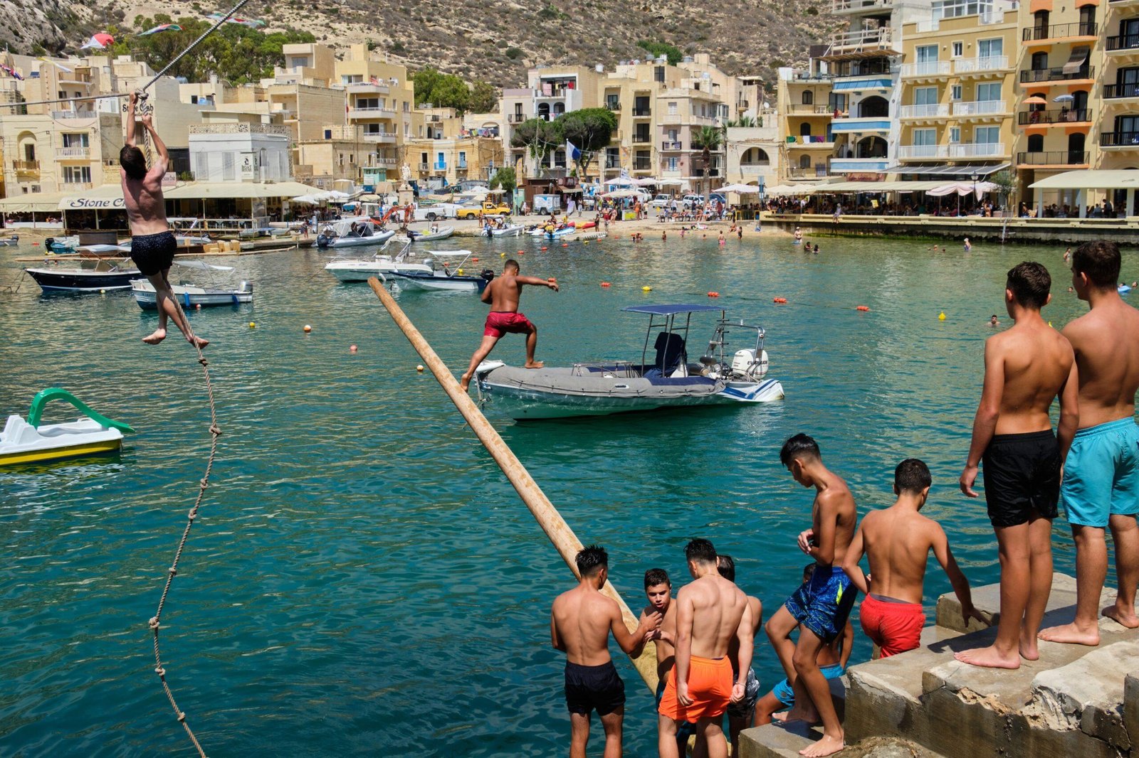 Men and boys in swimming shorts gather on the edge of Xlendi Bay by a greasy pole that juts out over the water.