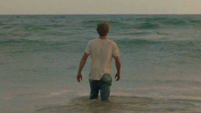 A man, wearing jeans and a white t-shirt, is wading out to sea.