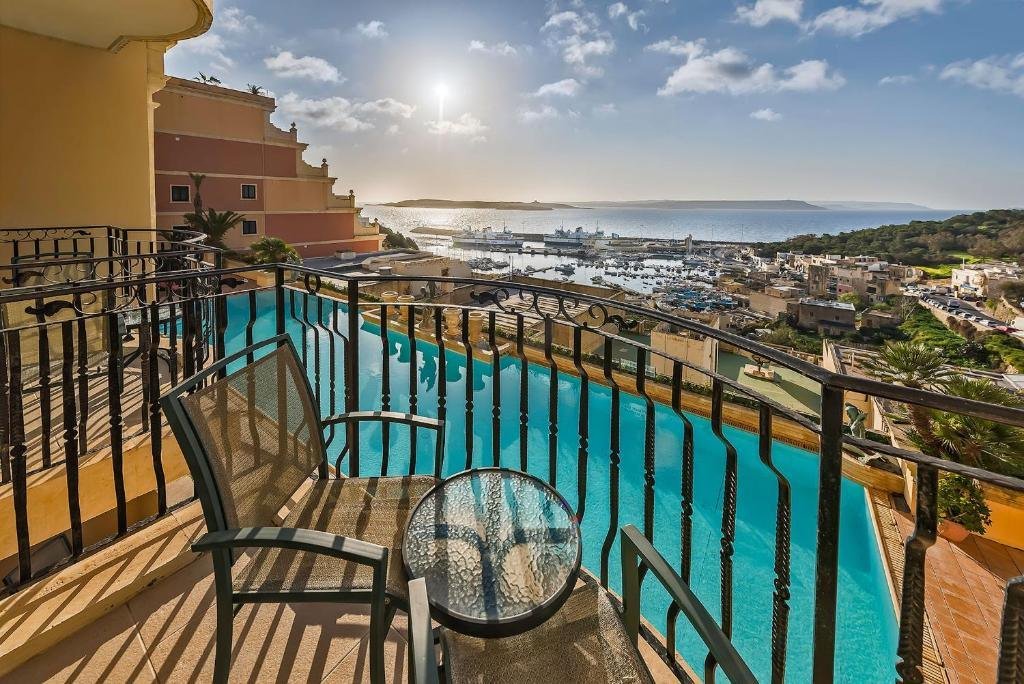 A hotel balcony with two chairs and table, overlooking a swimming pool and Mgarr harbour.