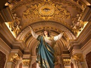 Statue of Our Lady, holding her hands aloft, inside an ornate church.