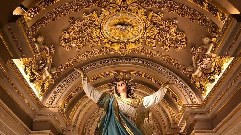 Statue of Our Lady, holding her hands aloft, inside an ornate church.