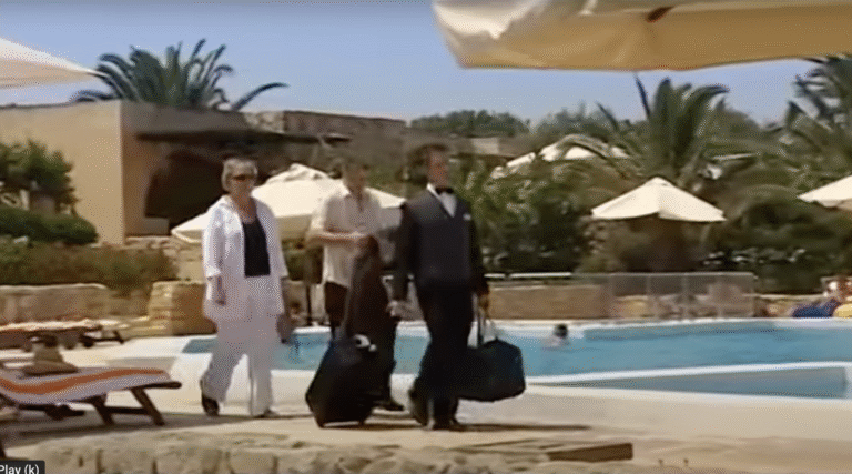 Two men and a woman walking by a hotel swimming pool, dragging suitcases.