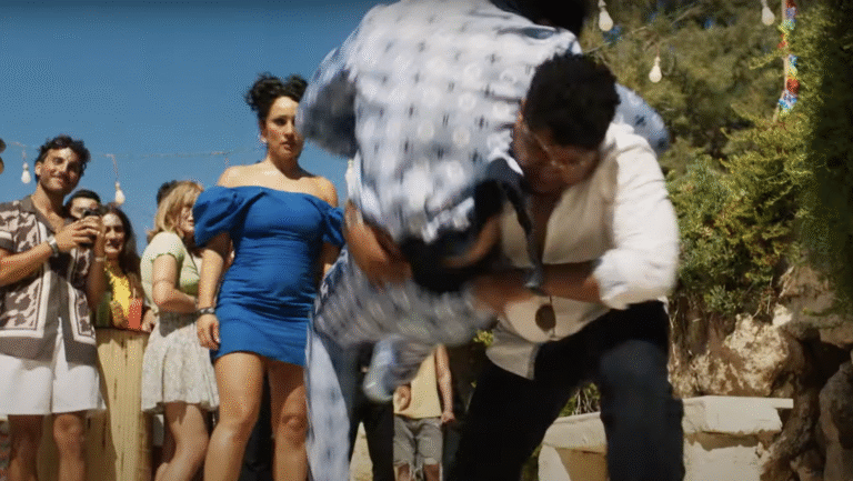 A man in a white shirt and jeans, rugby tackles a man in a plaid shirt as a woman in a short blue dress looks on.