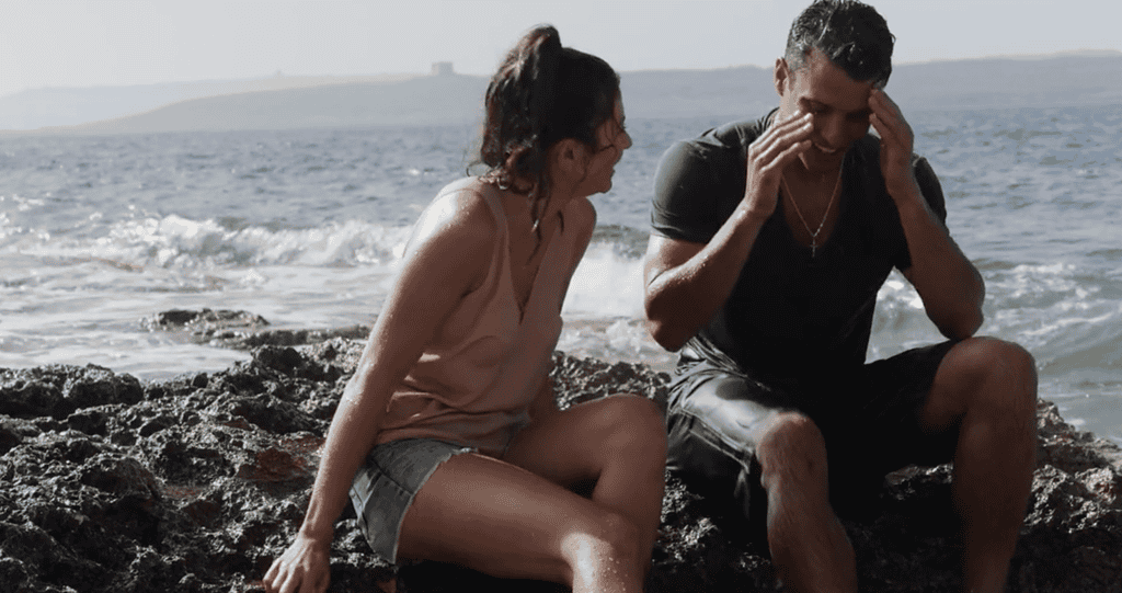 A man and woman sit on a rock with their backs to the sea. They are both fully clothed and soaking wet.