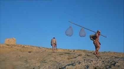 A man walks up a bare hill, another is walking out of sight to the right, carrying a long pole with fishing baskets.