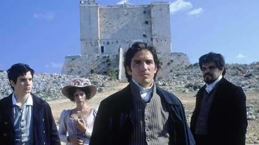 Three people, dressed as if from 18th century France, stand in front of a large, square, limestone tower.