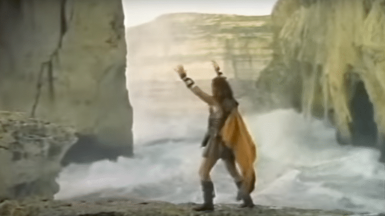A man, dressed as an ancient greek, stands with his legs and arms wide, on the sea shore.