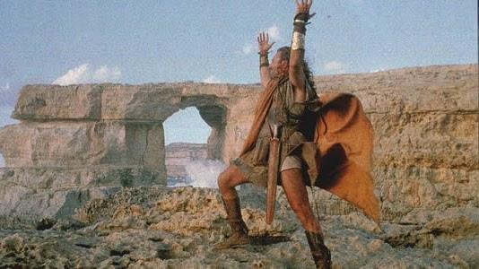 A man, dressed as an ancient greek, stands with his legs and arms wide, in front of the Azure window.
