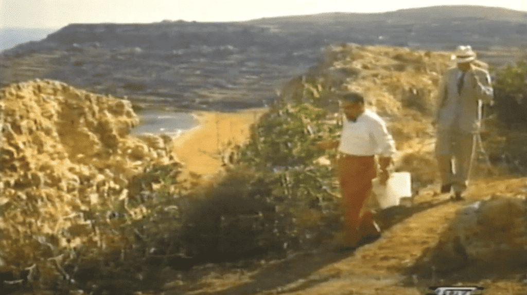 A man in a white shirt and red trousers walks along a hill, with a view of a sandy red beach.