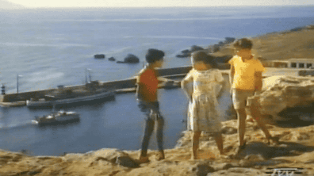 Three children stand talking on a hill, overlooking a harbour.