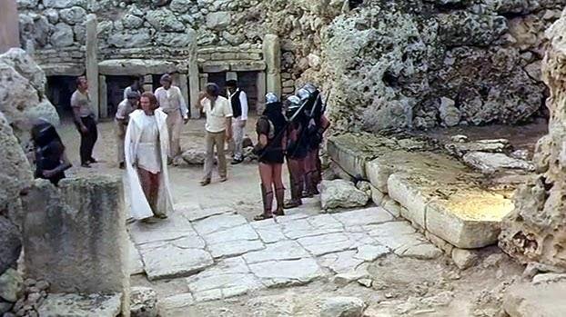 A group of people, some in long white robes, stand in front of the Ġgantija Temples.