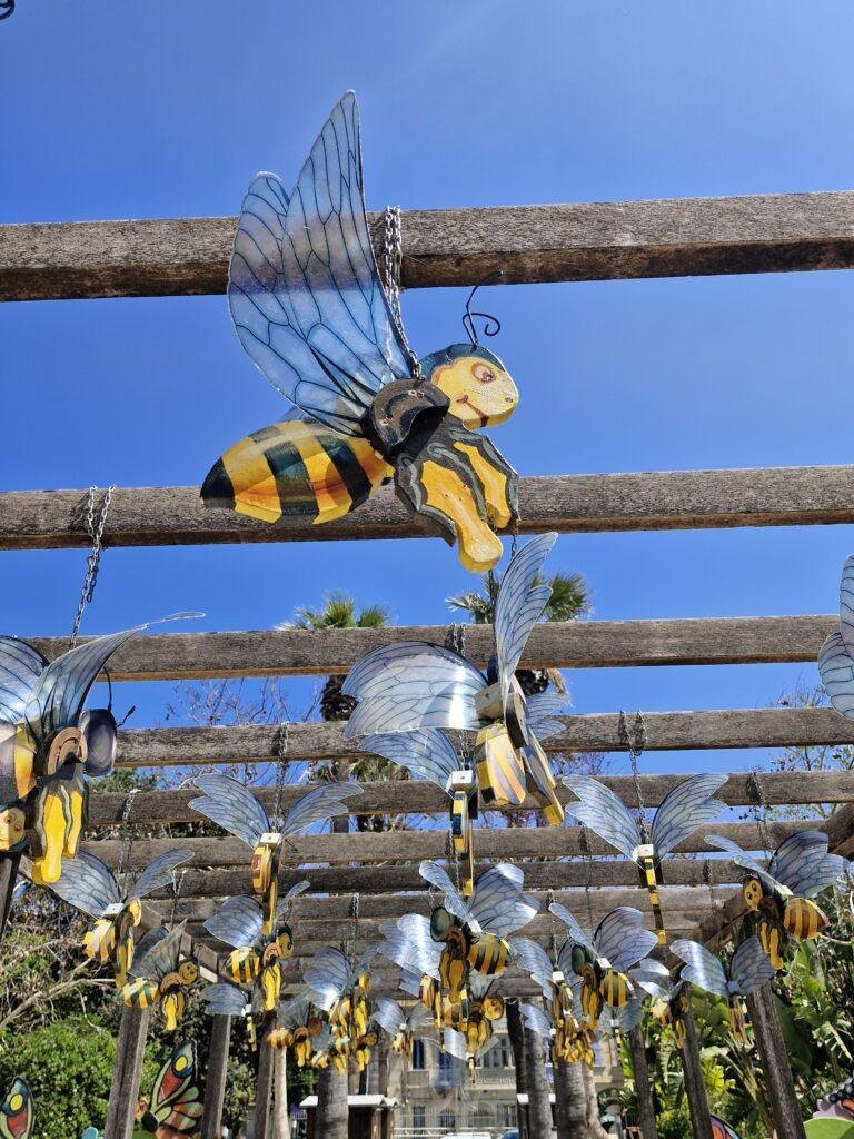 A wooden caricature of a honey bee hangs from a cross-beam against a blue sky. There are more wooden honey bees in the background.