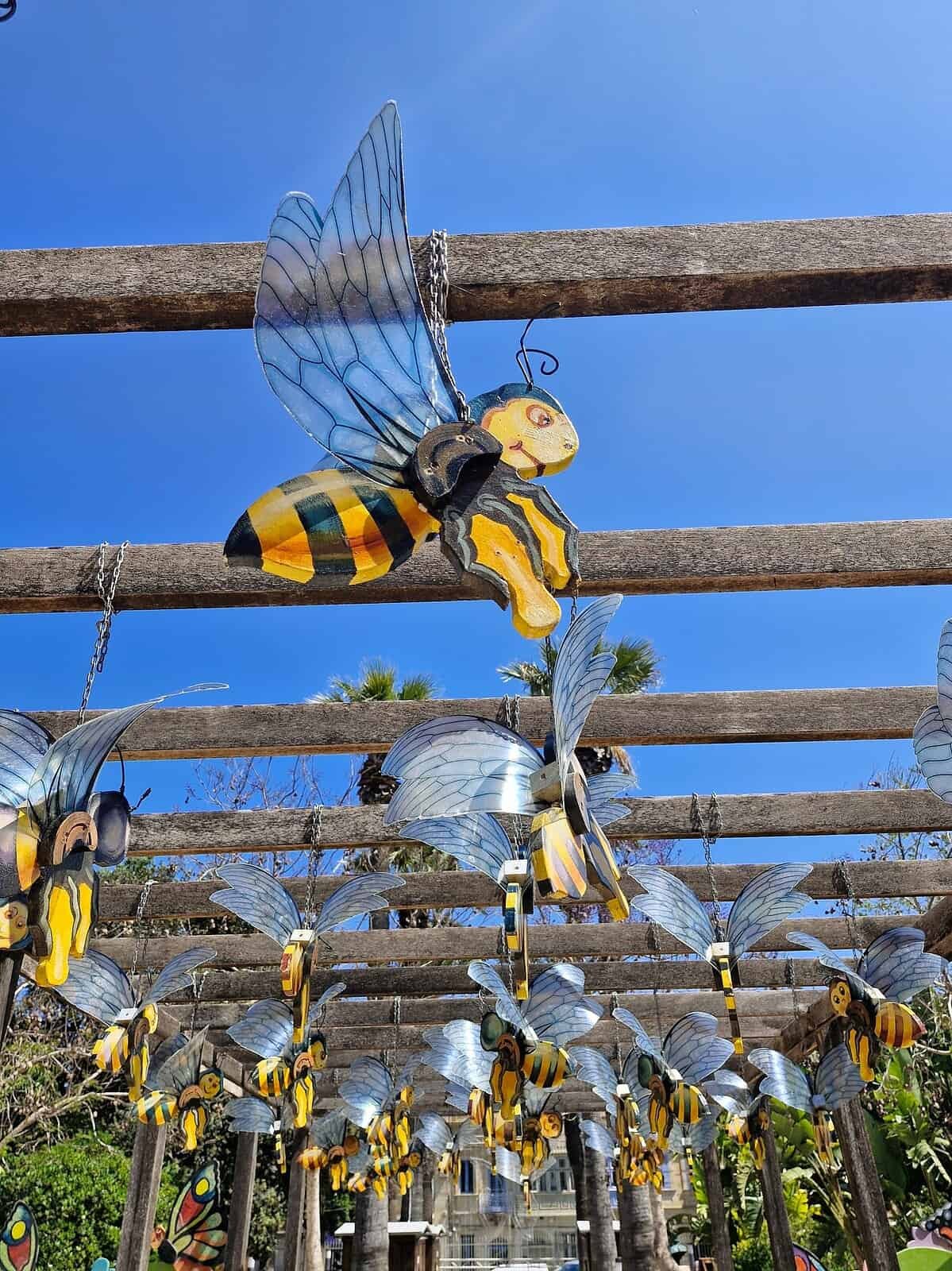 A wooden caricature of a honey bee hangs from a cross-beam against a blue sky. There are more wooden honey bees in the background.