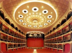 A wide shot of an opulent Opera House taken from the rear of the auditorium. The red chairs of the stalls stretch out in front, with three rows of boxes lining either side.