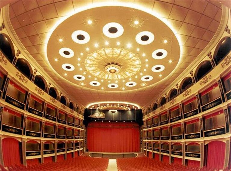 A wide shot of an opulent Opera House taken from the rear of the auditorium. The red chairs of the stalls stretch out in front, with three rows of boxes lining either side.