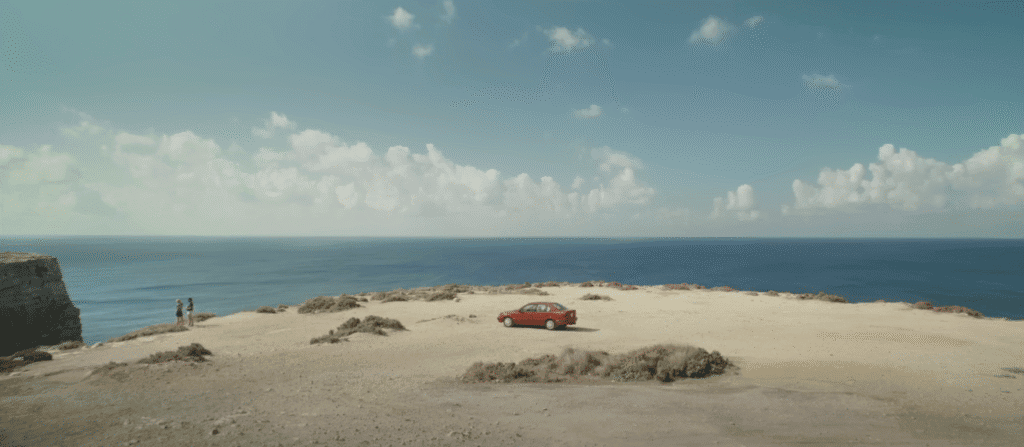 A red car parked on a limestone plateau overlooking the sea.