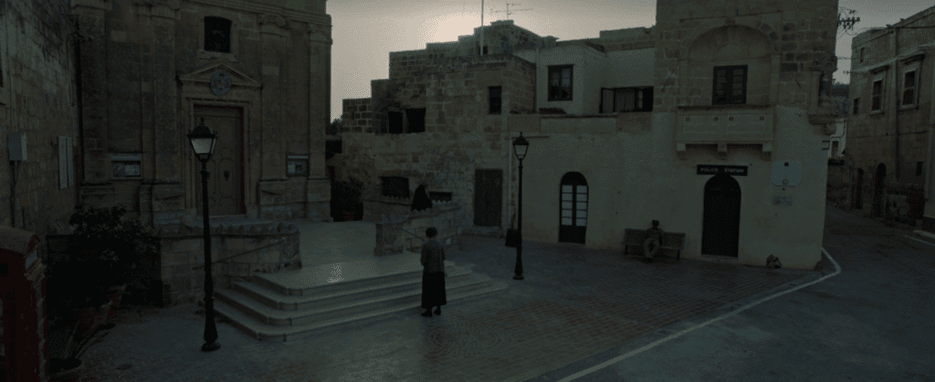 A lone female figure stands in a small village square..