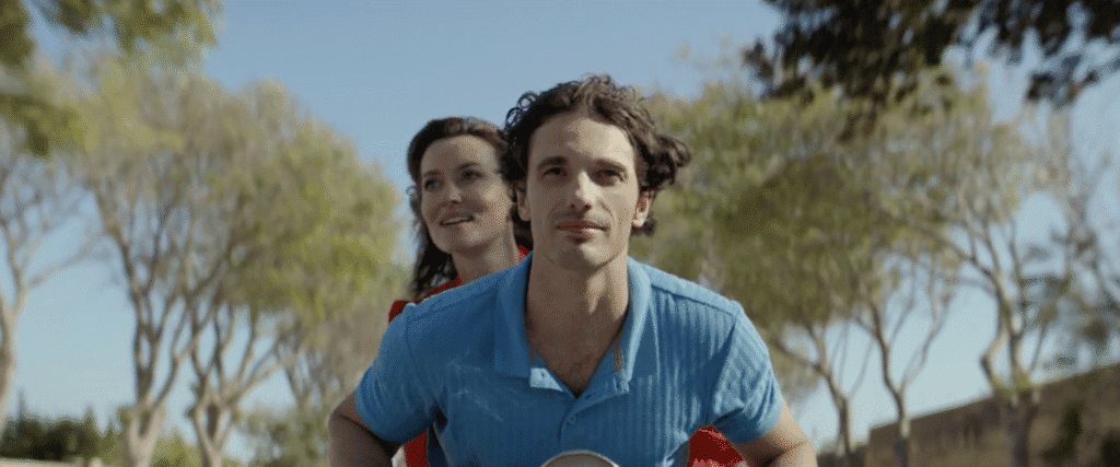 A woman in a flowing red dress rides behind a young man on a scooter down a country road.