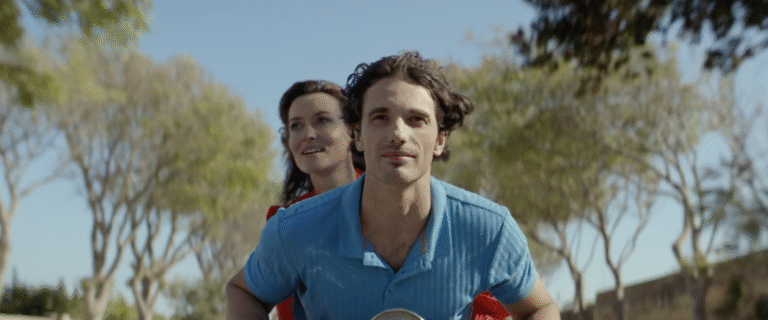 A woman in a flowing red dress rides behind a young man on a scooter down a country road.