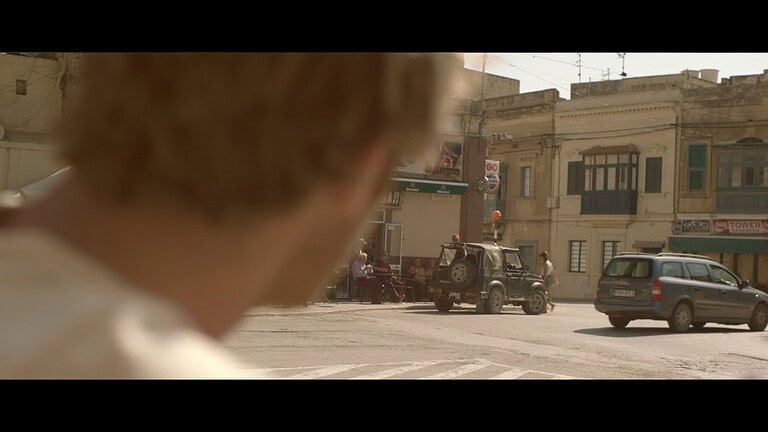 A man's head fills the left-hand side of the screen as looks across a road.