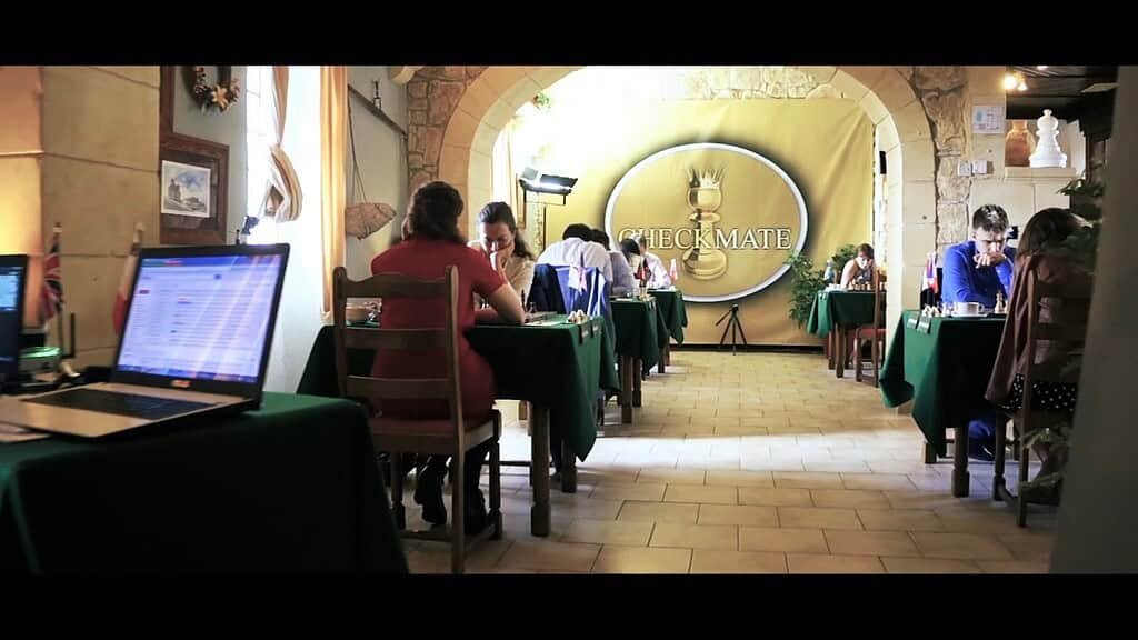 A large room, with limestone walls and a tiled floor. There are small tables down each side with green table clothes. Two people sit at each table with a chess board between them.