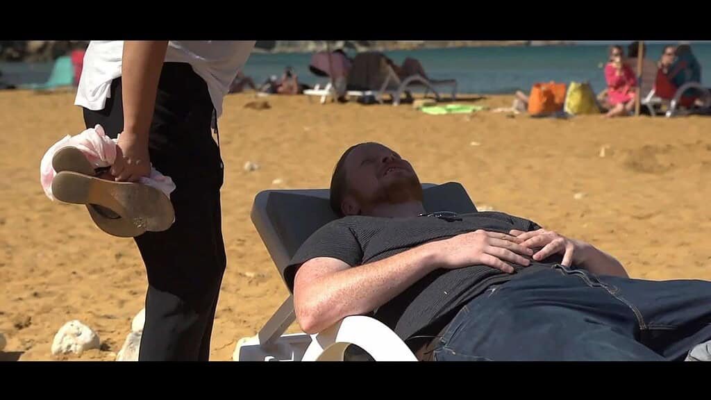 A woman stands over a man lying on a sun lounger on a sandy beach, her shadow is falling over his face.