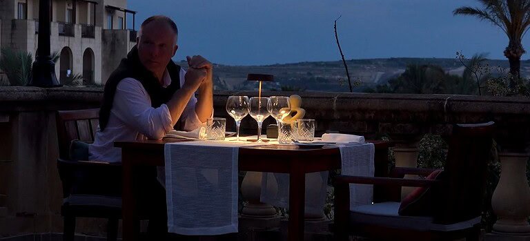 A man sits a two-person dining table at dusk. The table is lit by a lamp and has a white runner and several empty glasses.