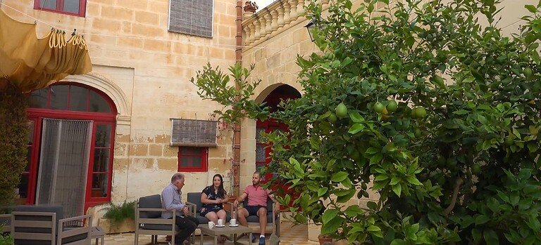 Two men and a woman sit on wooden chairs in the courtyard of a limestone house.