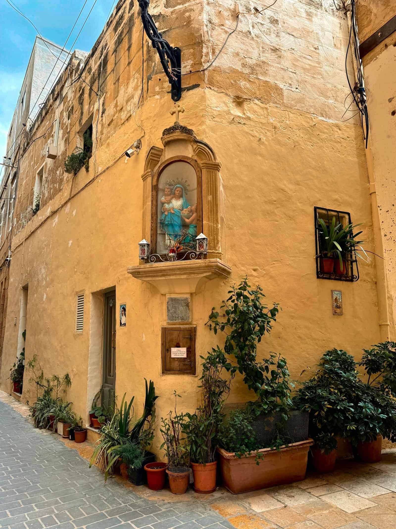 Charming corner house in Malta featuring a classic religious icon and vibrant potted plants.