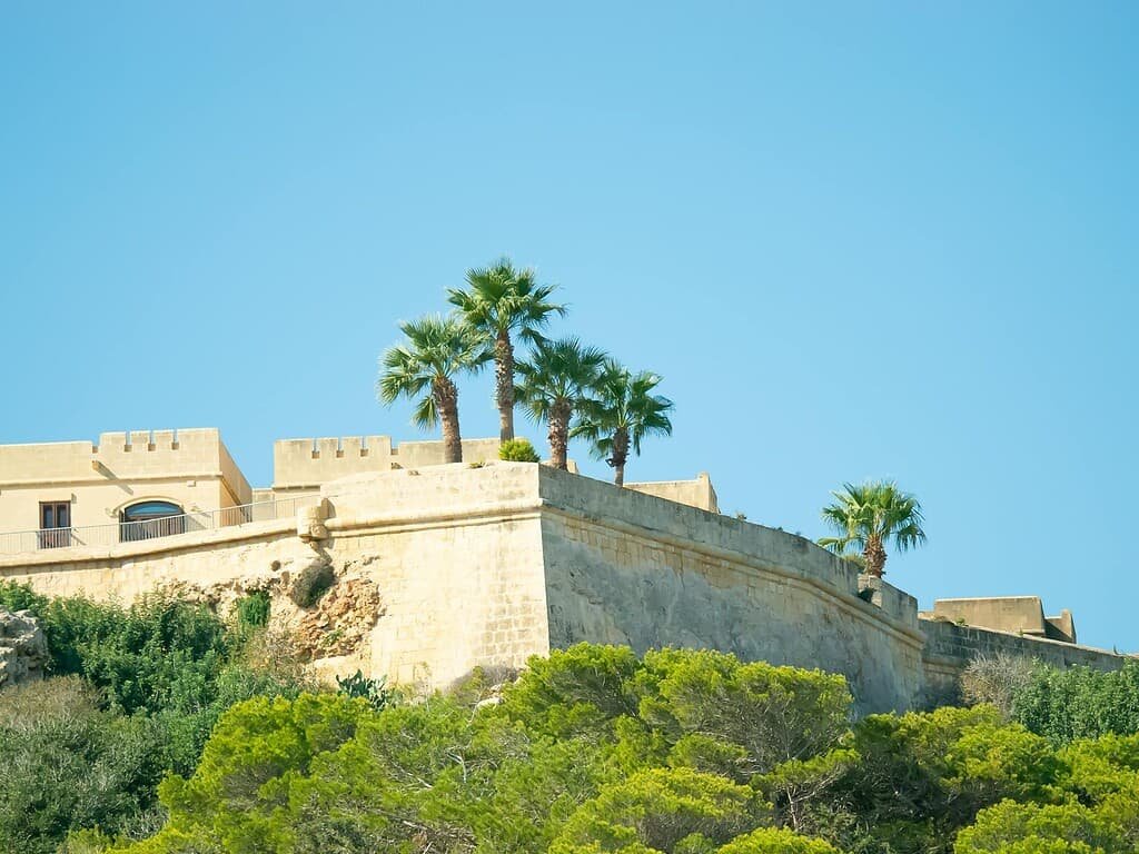 Explore the stunning architecture of an ancient fortress in sunny Malta with palm trees and clear blue skies.