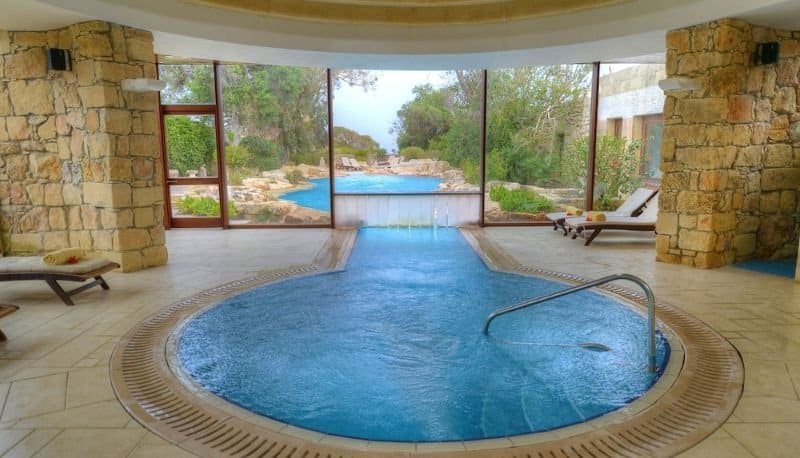 Large indoor jacuzzi  at the Ta' Cenc resort, set in a spacious limestone room, the jacuzzi  has a channel that leads to an outdoor pool, framed by trees.